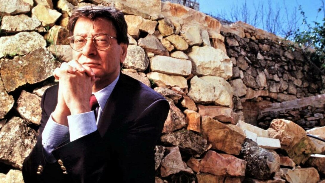 A photograph of the famous Palestinian poet Mahmoud Darwish. He is wearing a blue sweater, sitting with his hands below his chin, looking contemplative. He is in front of a brown stone wall in Palestine.